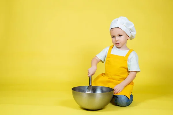小高加索男孩玩大厨 男孩穿围裙 戴着大厨帽坐在地板上 手里拿着碗 带着黄色背景的威士忌 — 图库照片