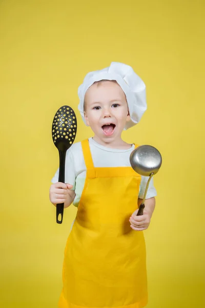 扮演厨师的小高加索男孩 戴着围裙的男孩和戴着厨师帽子的男孩拿着一只勺子和一个黄色背景的空勺子 — 图库照片