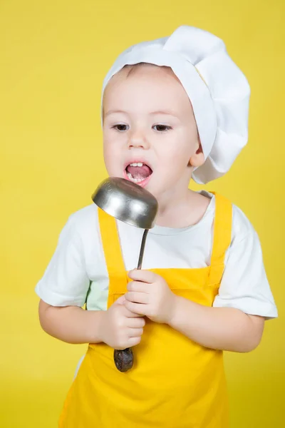 Piccolo caucasico ragazzo giocare chef, ragazzo in grembiule e cuochi cappello lecca il mestolo — Foto Stock