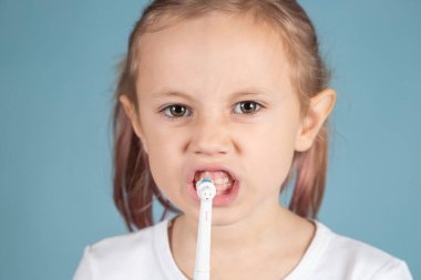 Gülümseyen küçük beyaz kız dişlerini fırçalıyor mavi arka planda duruyor, hijyen kavramı.