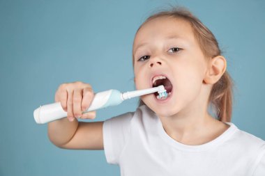 Gülümseyen küçük beyaz kız dişlerini fırçalıyor mavi arka planda duruyor, hijyen kavramı.