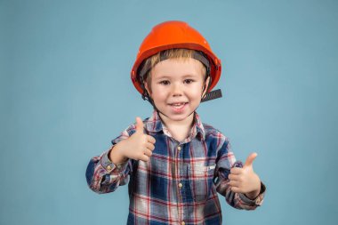 Mavi arka planda koruyucu turuncu kasklı şirin bir çocuğun portresi baş parmağını gösteriyor. İnşaat ve onarım kavramı.