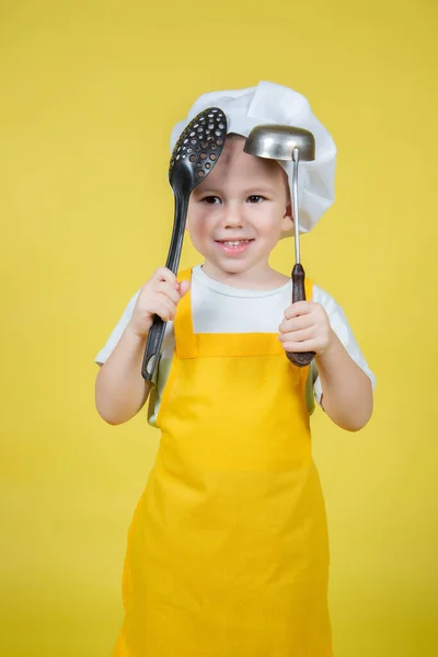 扮演厨师的小高加索男孩 戴着围裙的男孩和戴着厨师帽子的男孩拿着一只勺子和一个黄色背景的空勺子 — 图库照片