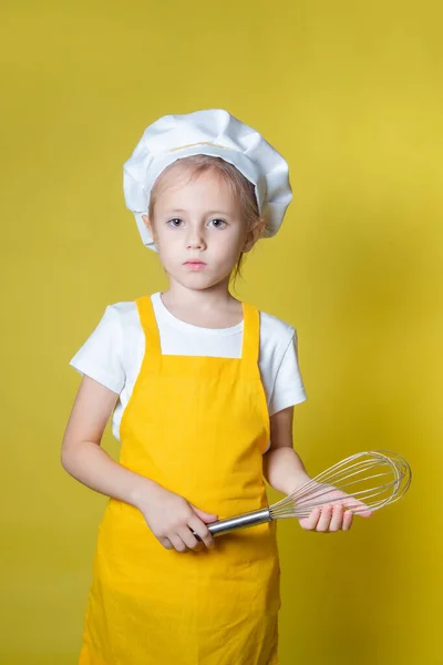 扮演厨师的小高加索女孩 戴着围裙的女孩 戴着厨师帽 拿着威士忌在黄色背景下鞭打奶油 — 图库照片