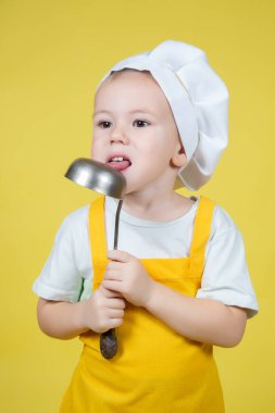 Küçük beyaz çocuk aşçıyı oynuyor, önlüklü çocuk ve şef şapkası sarı arka plandaki kepçeyi yalıyor.