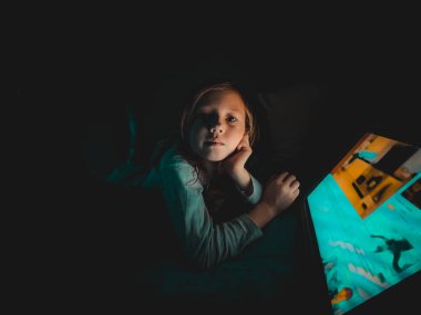 Küçük beyaz bir kız akşamleyin karanlıkta tablete bakıyor. Yatağın üstünde, evde yaşam tarzı fotoğrafçılığı yapıyor.