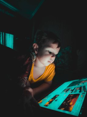 Küçük beyaz bir çocuk akşamleyin karanlıkta tablete bakıyor. Yatağın üstünde, evde yaşam tarzı fotoğrafçılığı yapıyor.