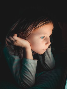 Küçük beyaz bir kız akşamleyin karanlıkta tablete bakıyor. Yatağın üstünde, evde yaşam tarzı fotoğrafçılığı yapıyor.