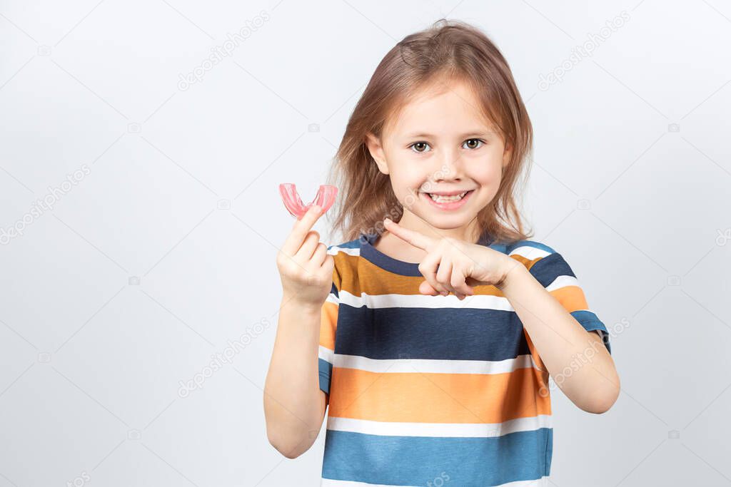 Cute little caucasian girl with blond hair is holding a pink dental myofunctional trainer on white the background. dental tariner is made to help equalize the growing teeth and correct bite