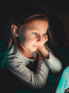 Küçük beyaz kız, akşamları karanlıkta tablete bakıyor. Yatağın üstünde, yaşam tarzı tonlu fotoğrafçılık, seçici odaklanma ve yumuşak odaklanma.