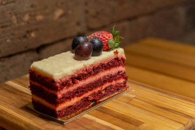 Üzümlü ve ahşap tepside çilekli Lezzetli Kırmızı Kadife kek ve arka planda kırsal tuğla duvar kompozisyonu. Bir dilim lezzetli ve güzel pasta. Seçici odak.