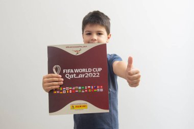 Bauru, Brezilya - 12.09.22: Gülümseyen ve elinde Katar 2022 dünya kupası albümü olan tatlı bir çocuğun ön görünümü - FIFA ve başparmaklar yukarı. Kupa çıkartmaları dünyanın her yerinden insanlar tarafından toplanıyor. Seçici odak.