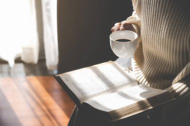 Asyalı kadın elinde bir fincan kahve tutuyor ve sabah kalktıktan sonra pencerenin yanında kitap okuyor. Sabah yaşam tarzı konsepti.