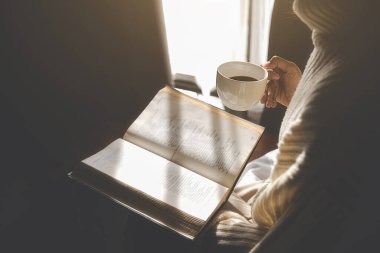 Asyalı kadın elinde bir fincan kahve tutuyor ve sabah kalktıktan sonra pencerenin yanında kitap okuyor. Sabah yaşam tarzı konsepti.