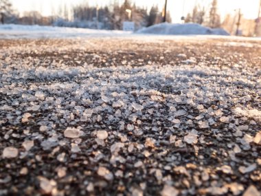 Salt grains on icy sidewalk surface in the winter. Applying salt to keep road clear and people safe in winter weather from ice or snow. Macro view of salt grains in sunlight in winter clipart