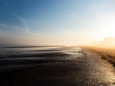 Güneşin doğuşuyla birlikte, sisli ve sisli bir sabahın erken saatlerinde, güzel, huzurlu Baltık deniz sahili manzarası. Islak kum, ışığı yansıtıyor ve havada beyaz bir sis var.