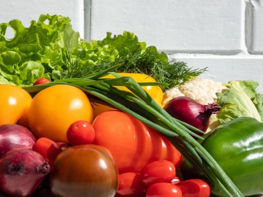 Bir yığın taze, sağlıklı ve renkli sebzeler parlak güneş ışığı altında ve arka planda beyaz tuğla bir duvar var. Domates, soğan, havuç, biber, karnabahar, salata, dereotu ve yeşiller masada.