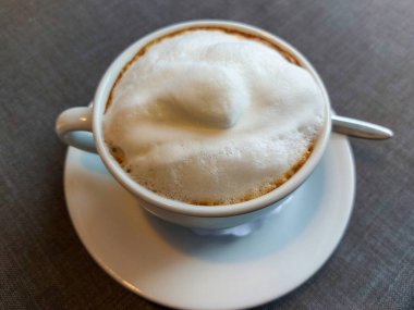 Pencerenin önündeki nötr gri masa örtüsüyle kapuçino kaplı beyaz süt köpüklü kahve kupasının görüntüsü. Sıcak içecek.