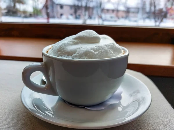 窓の前にある中性灰色のテーブルクロスで覆われたテーブルの上に ふわふわの白いミルクの入ったカプチーノコーヒーのマグカップの眺め ホットドリンク — ストック写真