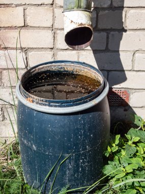 Big, blue plastic water barrel reused for collecting and storing rainwater from roof run-off in urban city environment with brick wall, asphalt and metal gate in the frame