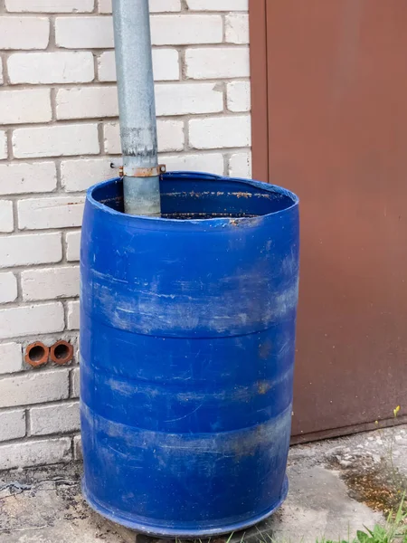 Big, blue plastic water barrel reused for collecting and storing rainwater from roof run-off in urban city environment with brick wall, asphalt and metal gate in the frame