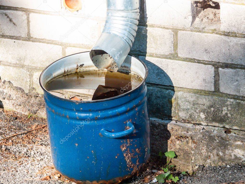 Big, blue metal pot reused outdoors for collecting and storing rainwater from roof run-off in urban city environment with brick wall and asphalt in the frame. Rainwater harvesting