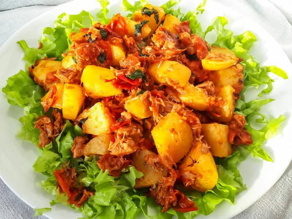 Close-up of a hot meal of boiled potatoes prepared with olive oil, tuna and cherry tomatoes on green salad leaves on white plate in bright sunlight. Healthy meal