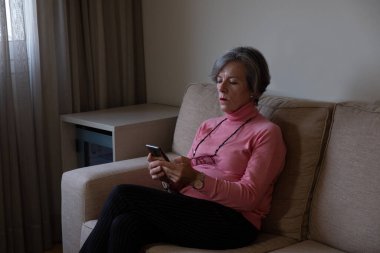 Olgun bir kadın koltukta oturmuş cep telefonunu kontrol ediyor.