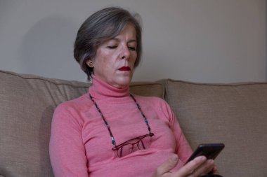 Yetişkin bir kadın, gözlüğü boynunda üç bacaklı bir sandalyede otururken cep telefonuna danışıyor..