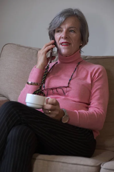 Mutlu olgun bir kadın koltukta oturup kahve içerken cep telefonuyla konuşuyor.