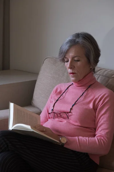 Gözlüklü yaşlı kadın kanepede dinleniyor ve kitap okuyor. Hobi kavramı.