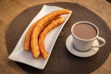 Damalı masa örtüsünün üzerinde bir fincan çikolatanın yanında churros tabağı..
