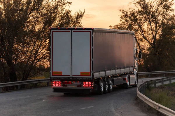 Truck with semi-trailer for general cargo seen from behind at sunset.