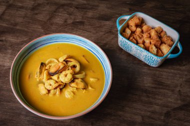 Deniz ürünleri çorbası, karides, midye, istiridye, hake ve mürekkep balığının yanında koyu ahşap bir masada kızarmış ekmek..