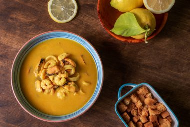 Karides, midye, istiridye, hake ve kalamar bazlı deniz ürünleri çorbası, koyu ahşap bir masada kızarmış ekmek ve limonla servis edilir..