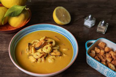 Karides, midye, istiridye, hake ve kalamar bazlı deniz ürünleri çorbası, koyu ahşap bir masada kızarmış ekmek ve limonla servis edilir..