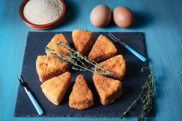 Presentation on a stone plate of fried camembert cheese together with the ingredients for the breading, breadcrumbs and eggs.