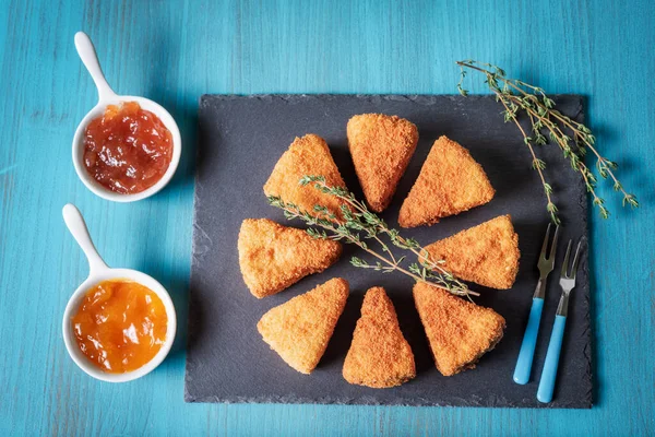 Presentation on a stone plate of fried camembert cheese next to jars with marmalade.