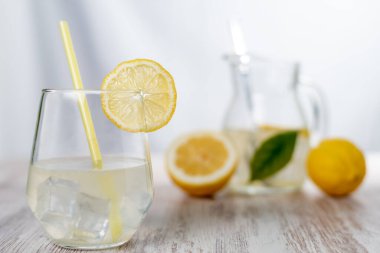 Limonata ve buzlu bardak, kenarında bir dilim limon, arka planda bir sürahi ve odak limonları..