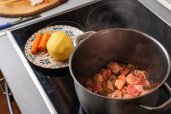 Pot over the fire of a ceramic hob cooking beef for a homemade stew, with potatoes and carrots prepared.