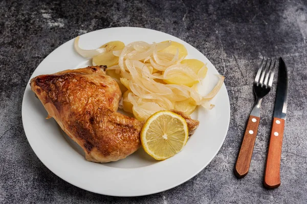 Lemon chicken dish, made in the home oven, with a thigh, potatoes, onion and a slice of lemon next to some cutlery.
