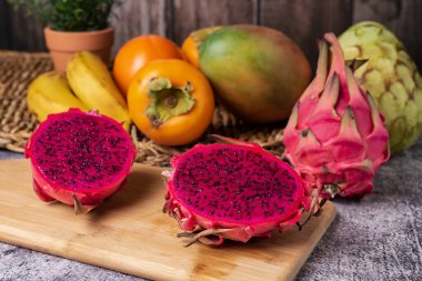 Pitahaya ya da ejderha meyvesi tahta bir tahtada ikiye ayrılır, pembe iç ve siyah tohumları ortaya çıkarır, arka planda Cherimoya, mango, hurma ve muz gibi bazı tropikal meyveler bulunur..