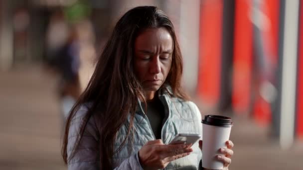 快適な大人の女性は、彼女の手の中にコーヒーカップで通りに立って、携帯電話にメッセージを書き込みます。ガジェットや都市生活の概念. — ストック動画