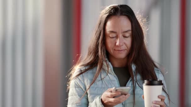 一位迷人的成年女性一边走路一边喝咖啡，一边在智能手机屏幕上打字。一个漂亮的女人在社交网络上交流，在街上消磨时光 — 图库视频影像