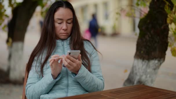 Взрослая женщина сидит за столом открытого кафе в ожидании заказа и смотрит на экран мобильного телефона — стоковое видео