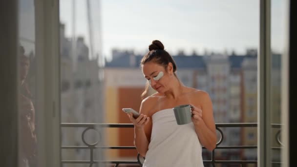 Una hermosa mujer está bebiendo café y usando su teléfono en un balcón en una ciudad europea. Desayuno en el balcón. Me despierto en la ciudad. El modo de vida europeo. — Vídeo de stock