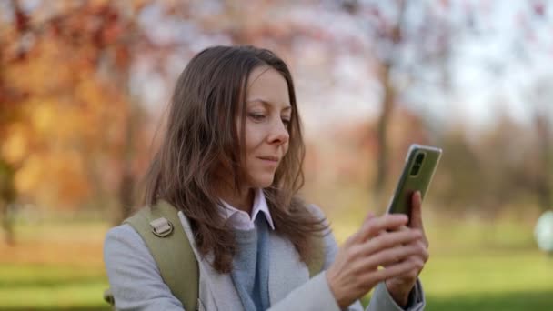 Sonbaharda şehir parkının ortasında internete erişmek için telefon kullanan yetişkin bir kadın turistin yakın çekimi. — Stok video