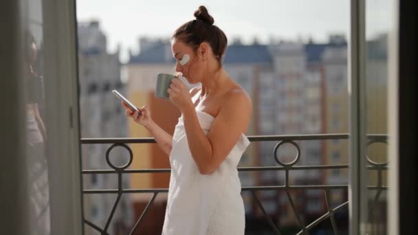 Una mujer adulta parada en un balcón con vistas a la ciudad y bebiendo una taza de té o café. Un medio de cuidado para el envejecimiento de la piel de la cara. Ver las noticias en la pantalla de un teléfono celular — Vídeo de stock
