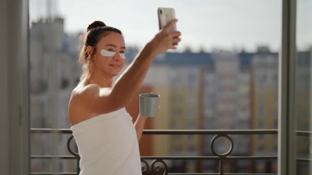 Una mujer adulta parada en un balcón con vistas a la ciudad y bebiendo una taza de té o café. Hace una foto selfie de sí mismo en el teléfono para compartir con los suscriptores buenos días. — Vídeos de Stock