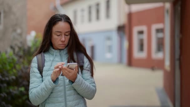Μια ενήλικη τουρίστρια εγκαθιστά έναν πλοηγό σε μια σύγχρονη τηλεφωνική συσκευή, περπατώντας στους δρόμους σε αστικές συνθήκες. ο ταξιδιώτης κάνει μια βόλτα και δημοσιεύει μια νέα ανάρτηση στην ιστοσελίδα — Αρχείο Βίντεο
